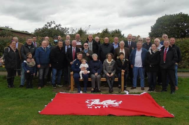 Some  of  Roy Davies’ family sitting on the new memorial bench, joined by members of Rugby Welsh, with the club flag in front. The bench is inscribed with the words ‘In memory of all those who loved our club - Rugby Welsh Rugby Football Club’