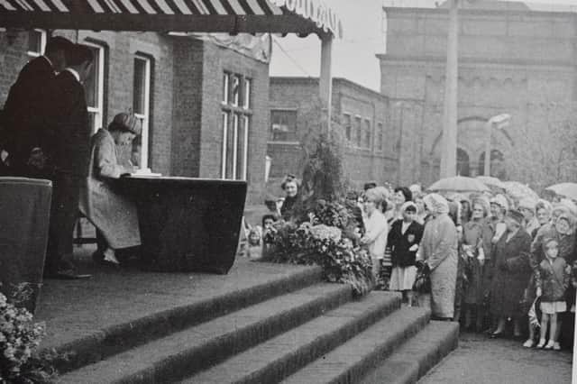 The Queen signs the works' visitor book as a crowd of Rugbeians watch.
